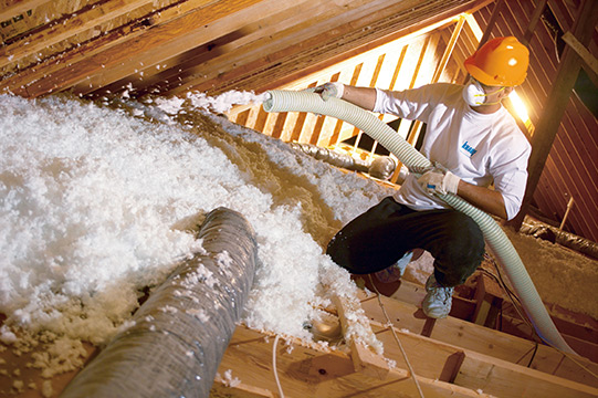 Technician wearing yellow hard hat, blowing loose-fill insulation into an unfinished attic.